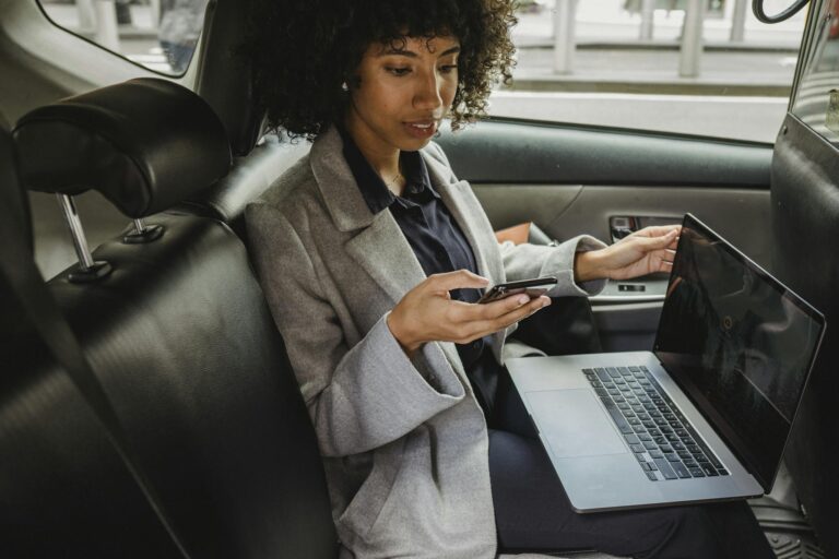 Crop African American female in formal clothes sitting with laptop on knees and browsing mobile phone in comfortable automobile