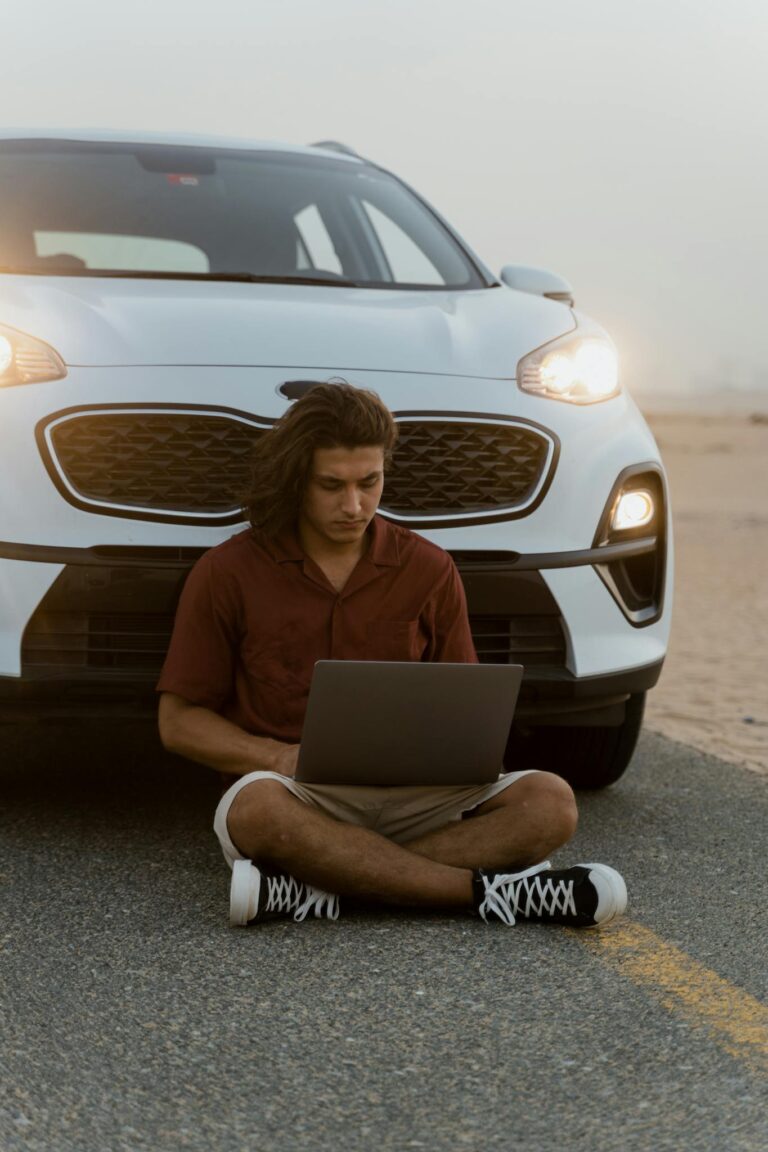 Man sitting with a laptop in front of a car on a desert road at dusk, enjoying a digital nomad lifestyle.