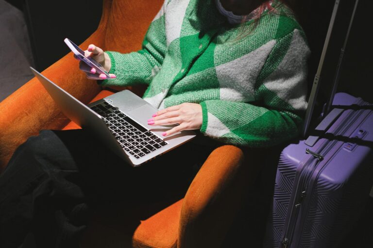 Woman in a knitted sweater working remotely on laptop with a smartphone, sitting in an armchair.